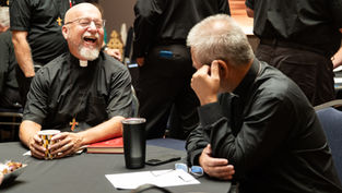 Two clergy in black shirts laugh at a table in a meeting room, with cups, papers, and others standing in the background.