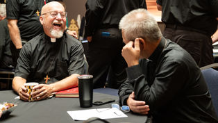 Two clergy in black shirts laugh at a table in a meeting room, with cups, papers, and others standing in the background.