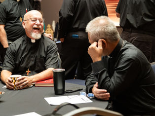 Two clergy in black shirts laugh at a table in a meeting room, with cups, papers, and others standing in the background.