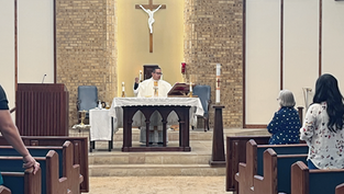 Priest in white vestments celebrates Mass at altar beneath crucifix, with congregants standing in pews of a brick church.