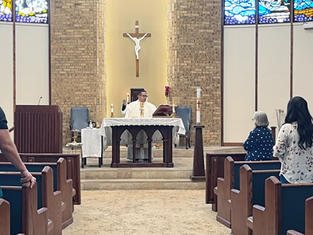 Priest in white vestments celebrates Mass at altar beneath crucifix, with congregants standing in pews of a brick church.