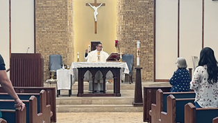 Priest in white vestments celebrates Mass at altar beneath crucifix, with congregants standing in pews of a brick church.