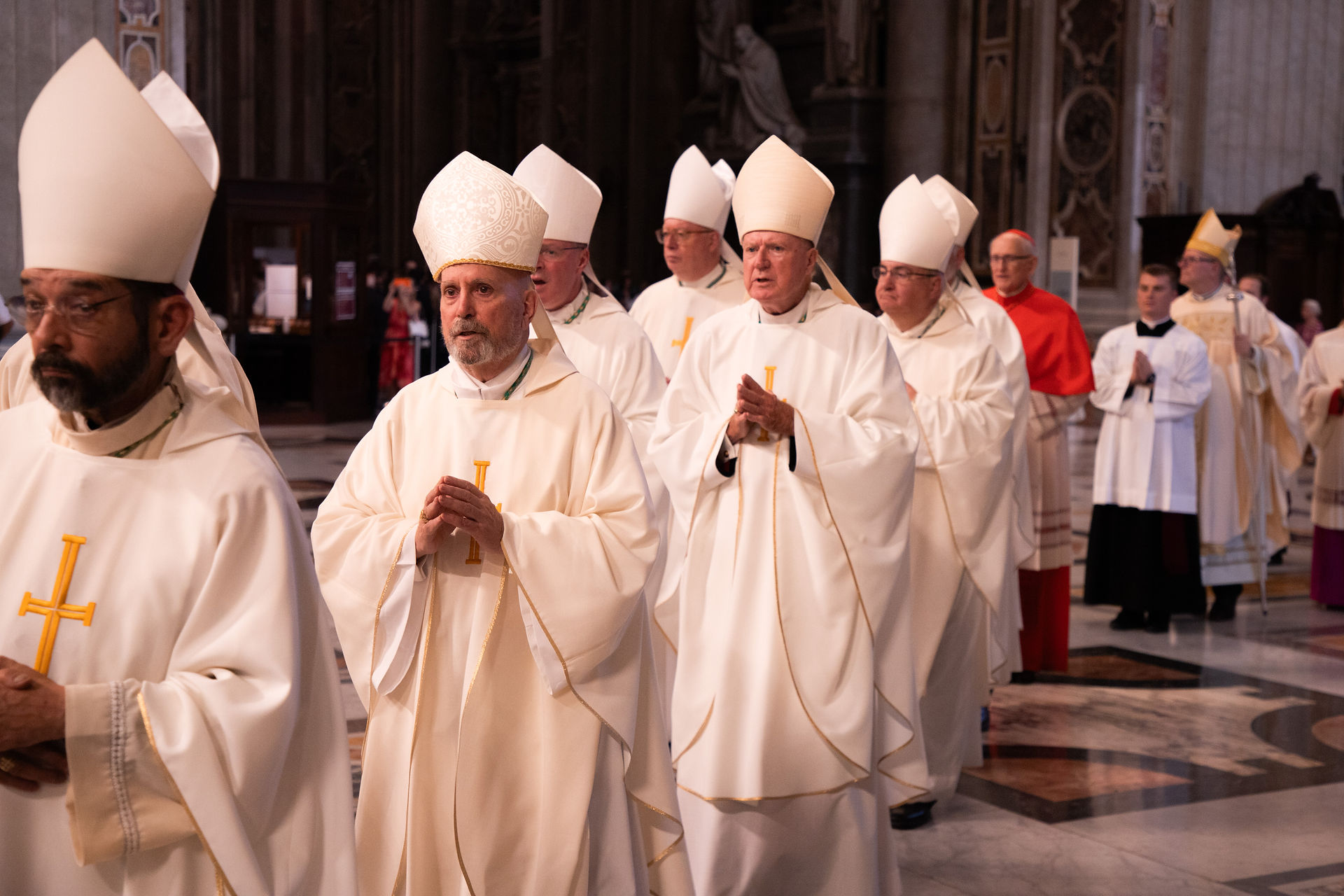 Bishops and clergy in white robes and miters process solemnly inside a cathedral, hands clasped in prayer.