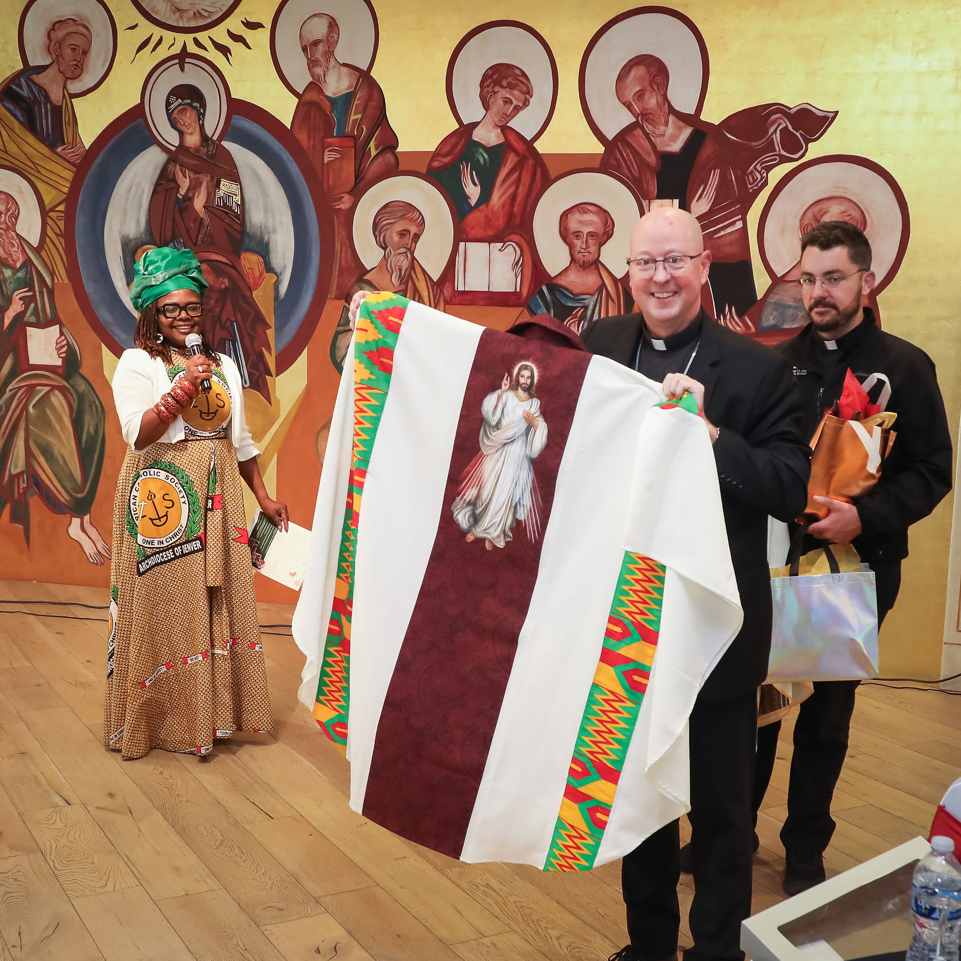 Smiling woman and two priests pose before a church mural, holding a white cloth with Jesus image and colorful trim.