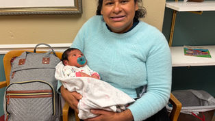 Woman in a light blue sweater cradles a baby with a pacifier in a cozy room, beside a gray bag and shelves with a toy bear.