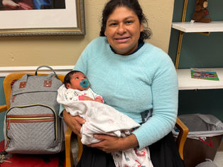 Woman in a light blue sweater cradles a baby with a pacifier in a cozy room, beside a gray bag and shelves with a toy bear.