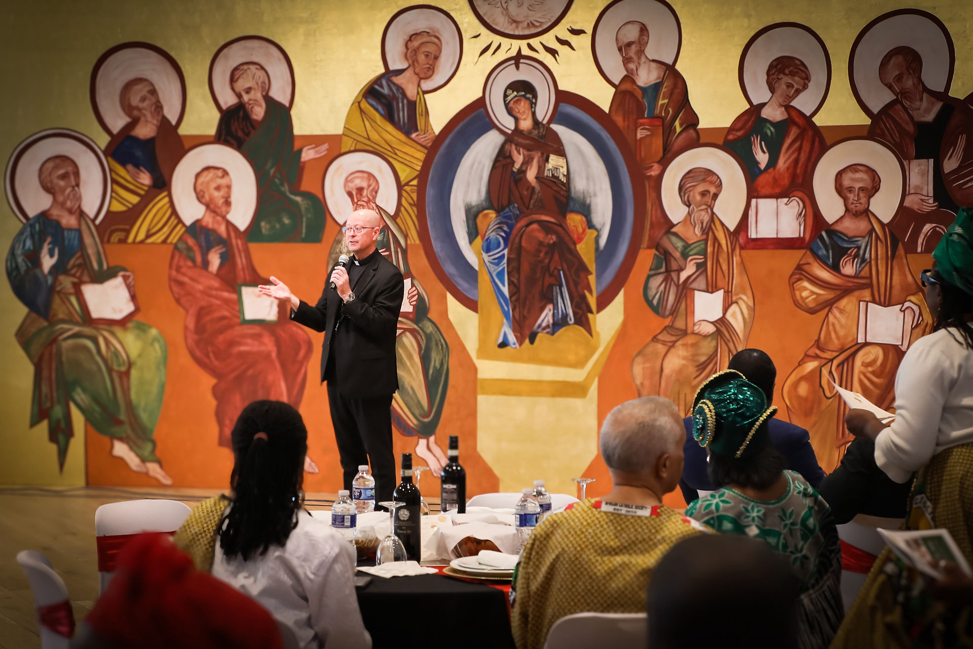 Priest speaks with microphone before seated audience in a hall with colorful saint mural; warm, reverent atmosphere.