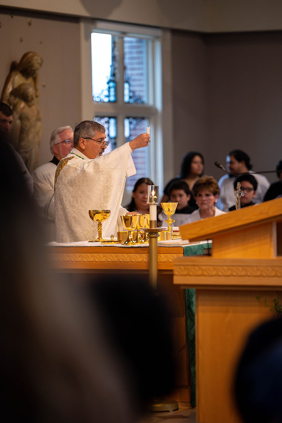 Priest in white vestments raises a communion wafer at a church altar with gold chalices as worshippers watch.