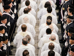 Clergy in white robes process between uniformed honor guards holding swords at a solemn indoor ceremony.