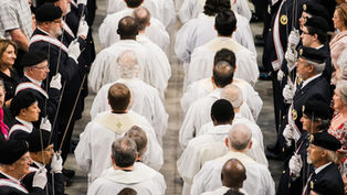 Clergy in white robes process between uniformed honor guards holding swords at a solemn indoor ceremony.