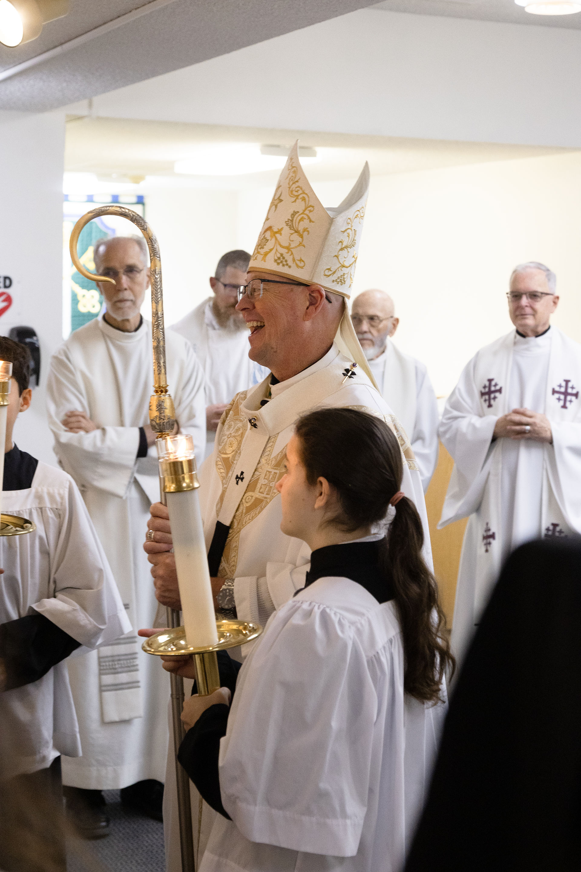 Smiling bishop in ornate mitre holds a lit candle in a church procession, surrounded by altar servers in white robes.