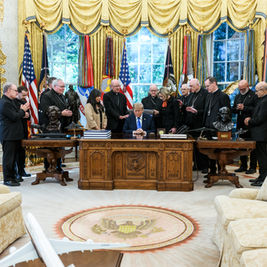 Trump sits at the Resolute Desk as clergy pray around him in the Oval Office, with flags and portraits behind.