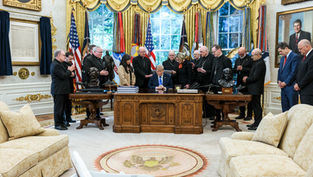 Trump sits at the Resolute Desk as clergy pray around him in the Oval Office, with flags and portraits behind.