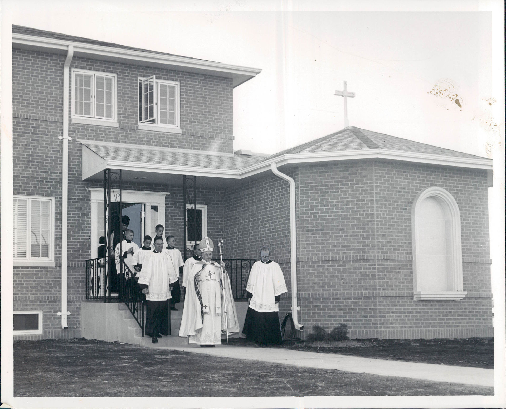 Bishop and clergy in vestments stand on the steps of a brick church, with a cross on the roof and a solemn mood.
