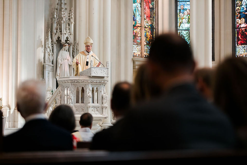 Bishop speaking from an ornate white pulpit in a cathedral, with blurred congregation in foreground and stained-glass windows behind