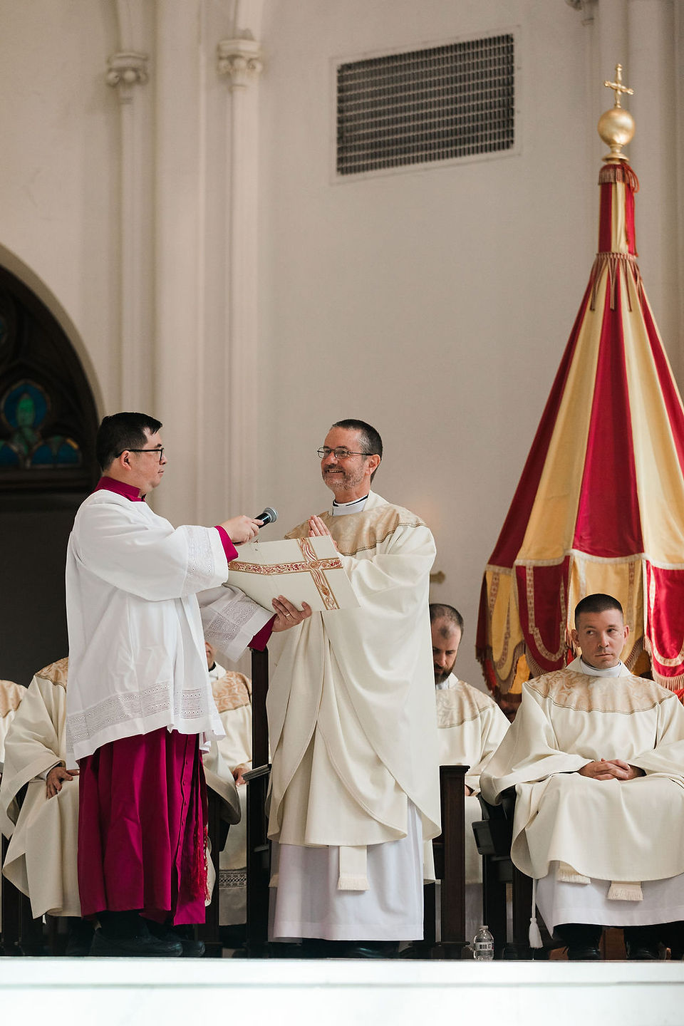 Priests in white robes share a liturgical book during a church ceremony, with a red-and-gold banner beside them.