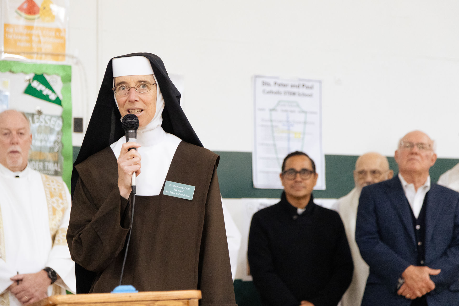 Nun in brown habit speaks into a microphone at a school event, with clergy and guests standing behind her in a bright hall