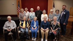 Smiling seniors and a priest pose in a church room with American and Christian flags.