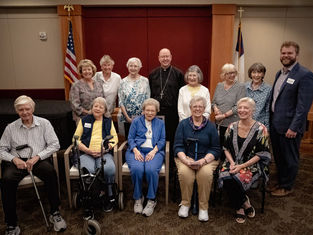 Smiling seniors and a priest pose in a church room with American and Christian flags.