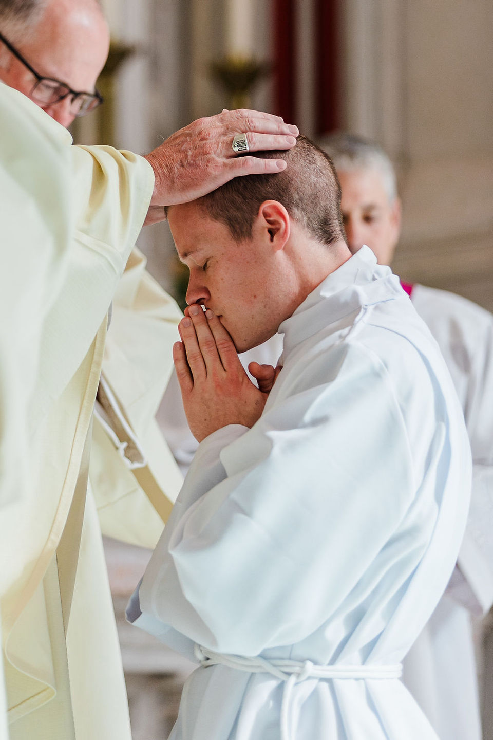 Bishop ordains a kneeling man in a white robe, touching his head as he prays in a softly lit church.