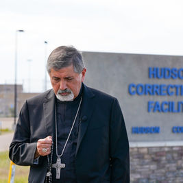 Cleric in black suit holds a rosary outside Hudson Correctional Facility sign in Hudson, Colorado, looking solemn.