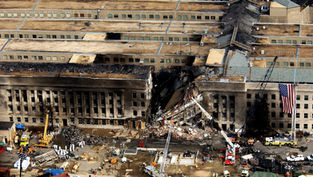 Burned Pentagon section with collapsed center, rescue crews and cranes below, American flag hanging on right, aftermath scene