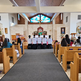 Children kneel in church pews during Mass before an altar with priest, crucifix, stained glass, and flowers.