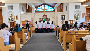 Children kneel in church pews during Mass before an altar with priest, crucifix, stained glass, and flowers.