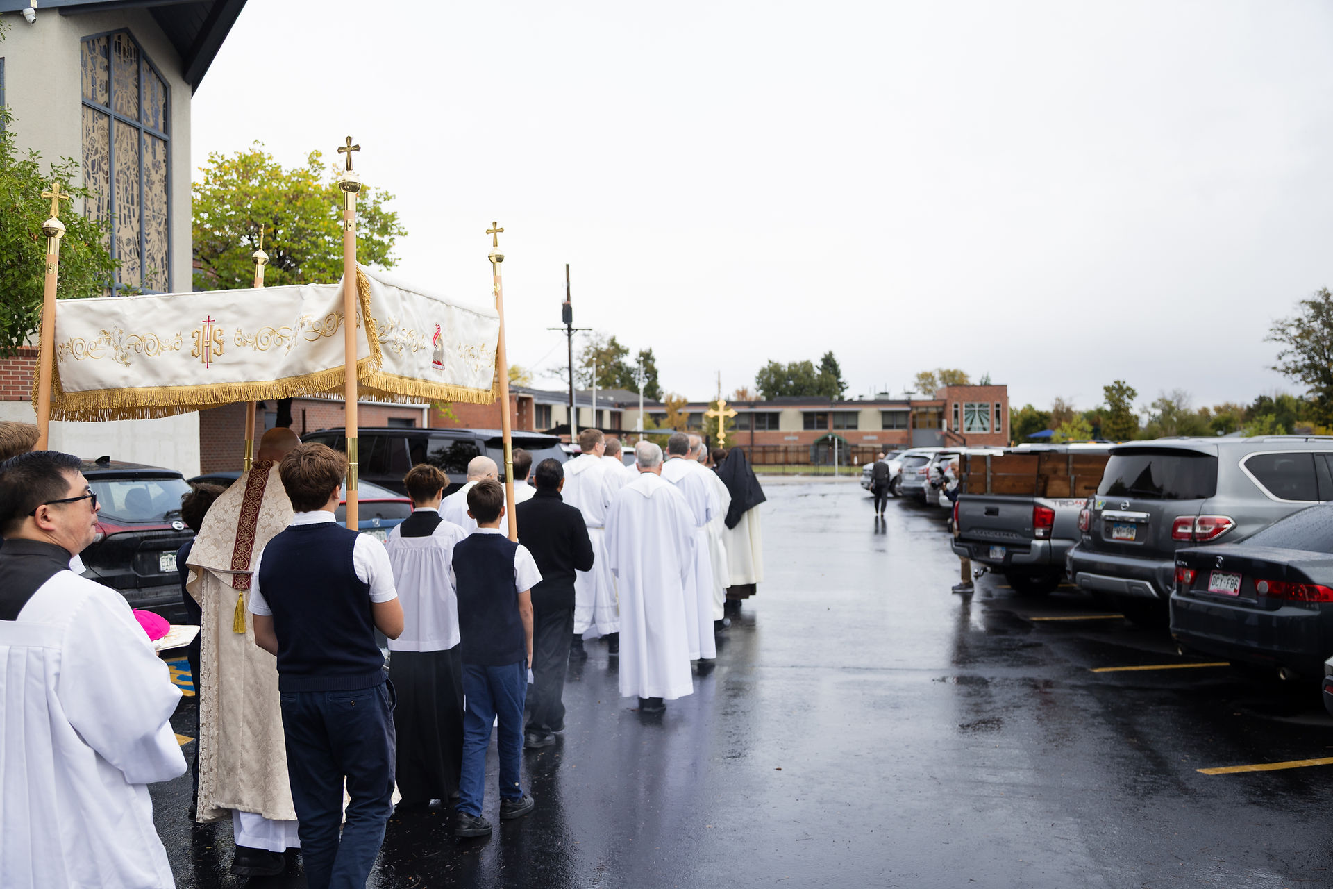 Clergy and boys in white procession under a gold-trimmed canopy in a rainy church parking lot, with parked cars.