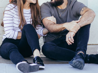 Couple sitting on a sidewalk, leaning together against a white wall, wearing casual black and striped clothes, looking affectionate.