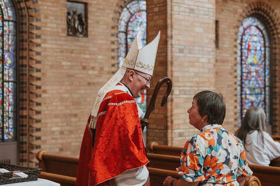 Bishop in red vestments and mitre chats with a woman in a colorful dress inside a church with stained-glass windows.
