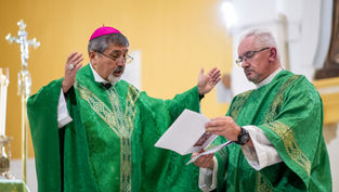 Two clergy in green vestments confer at the altar, one gesturing while the other reads from papers in a church.