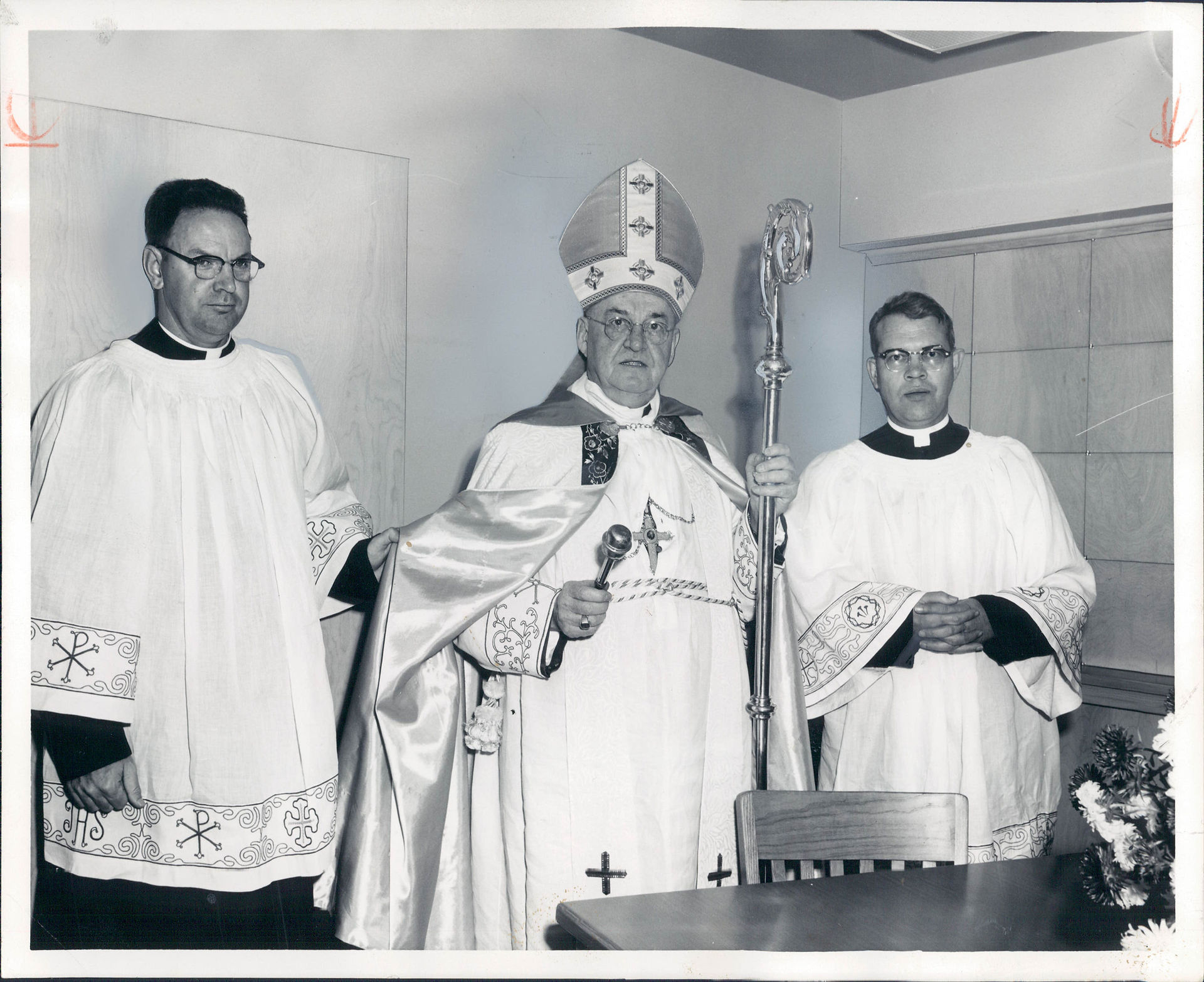 Three clergy in white vestments pose indoors; the bishop in a mitre holds a staff and microphone, solemn and formal.