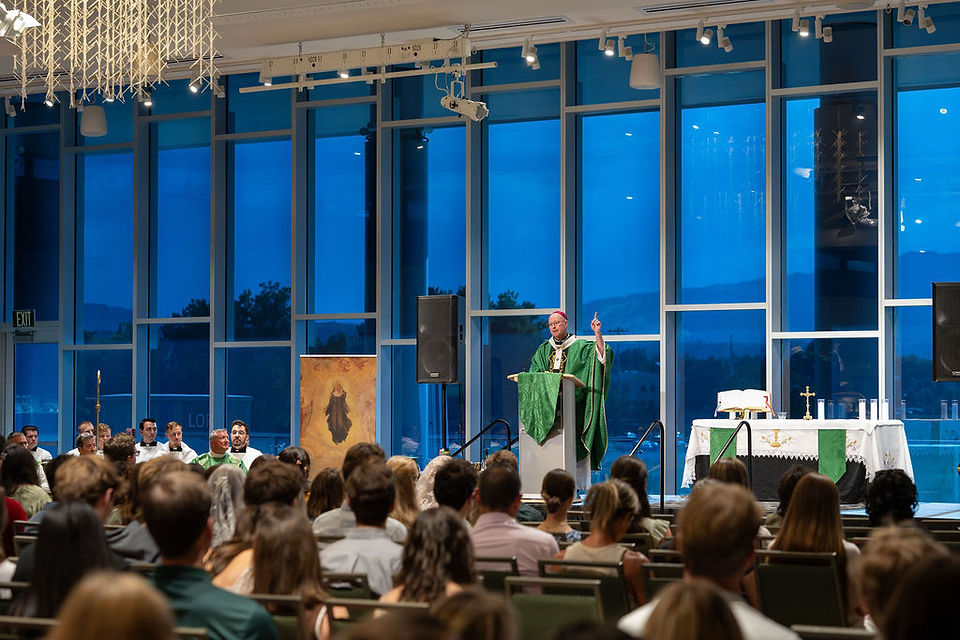 Priest in green vestments speaks at a podium to a seated congregation in a modern chapel with blue dusk windows and altar.