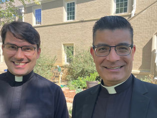 Two smiling priests in black clerical collars pose in a sunny courtyard outside a brick building with a statue.