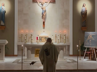 Deacon stands before a candlelit church altar with crucifix and saint statues, facing an easel painting in a quiet chapel.