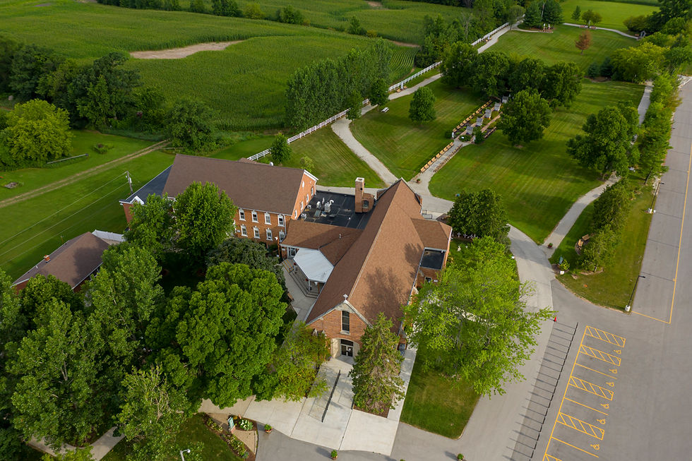 Aerial view of a brick building beside green fields, trees, and winding paths, with a parking lot marked for accessible spaces.