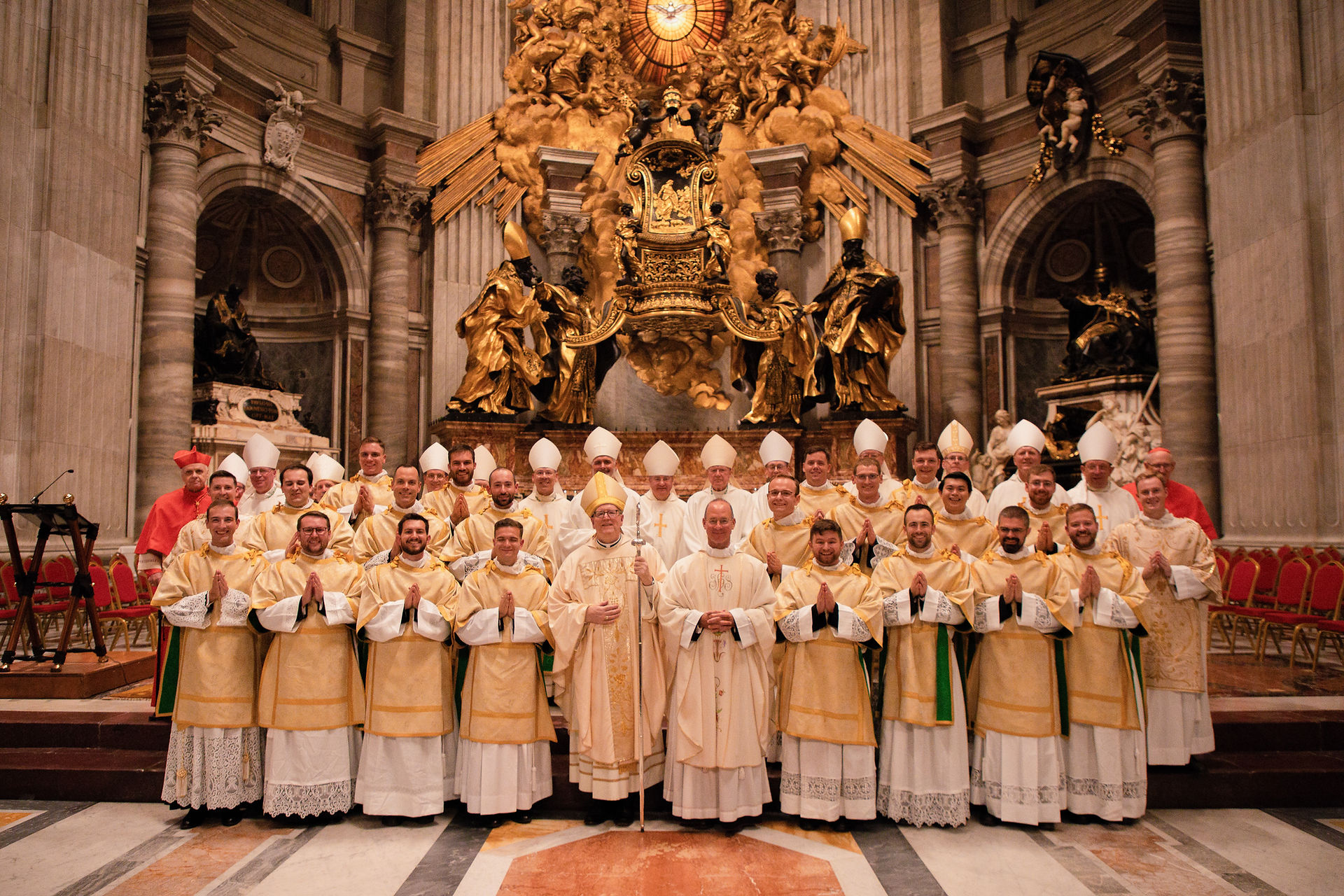 Group of Catholic clergy in gold and white vestments posing inside an ornate basilica before a gilded altar.