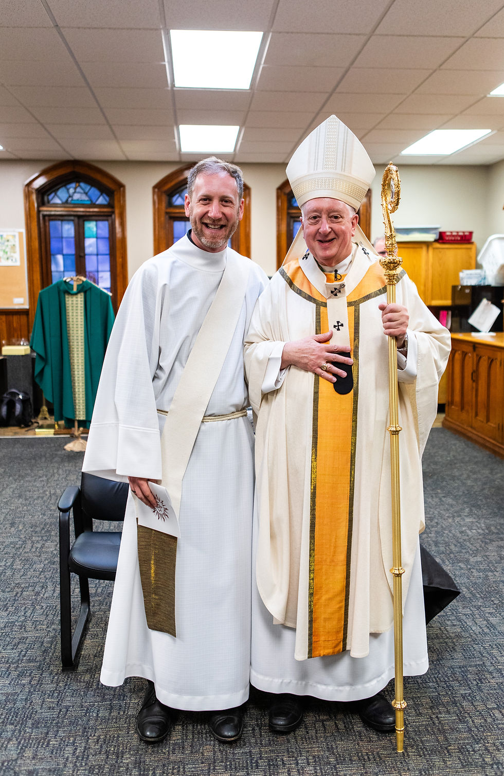 Smiling priest and bishop in white vestments pose in a church office, bishop holding a gold staff.