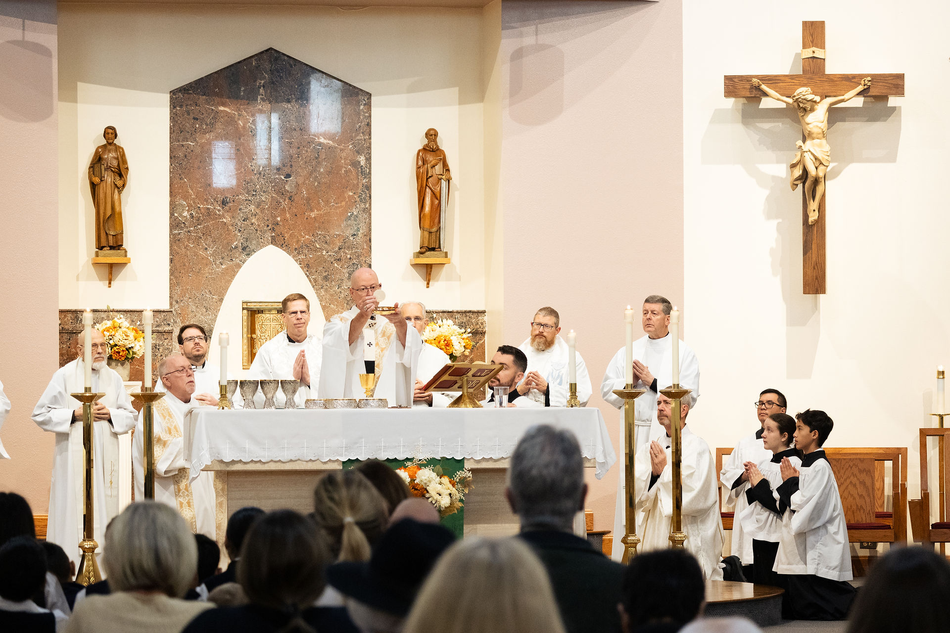 Priest elevates the Eucharist during a Catholic Mass, with clergy and altar servers around the altar beneath a large crucifix.