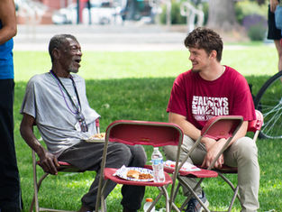 Two men chat and smile on red folding chairs at a park picnic, with plates of food and a water bottle; one shirt says ALWAYS MOVING.