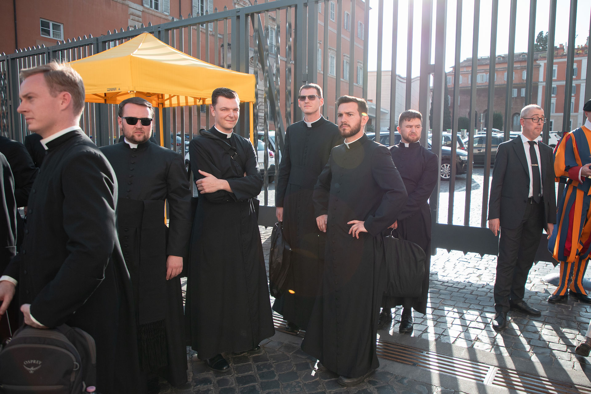 Group of Catholic seminarians in black cassocks stand outside a gated building, some in sunglasses, with a yellow canopy and bright sun.