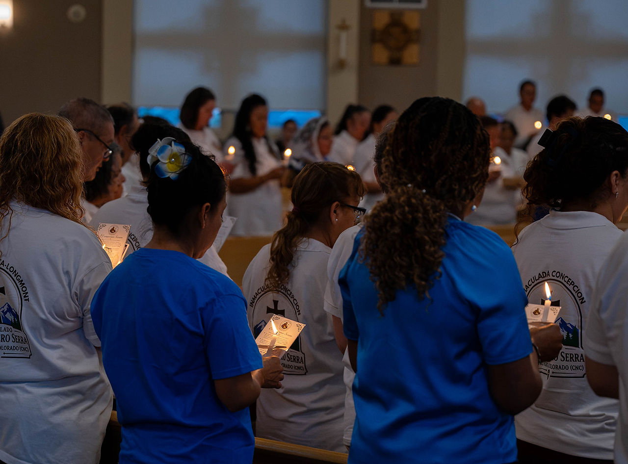 People in white and blue shirts hold lit candles during a candlelight church ceremony, with a solemn, prayerful mood.