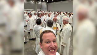 Smiling man takes a selfie in front of a large crowd of clergy in white robes inside a bright convention hall.
