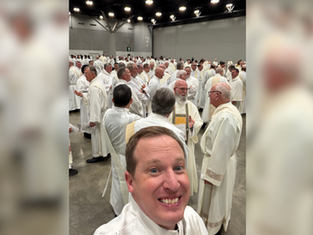 Smiling man takes a selfie in front of a large crowd of clergy in white robes inside a bright convention hall.