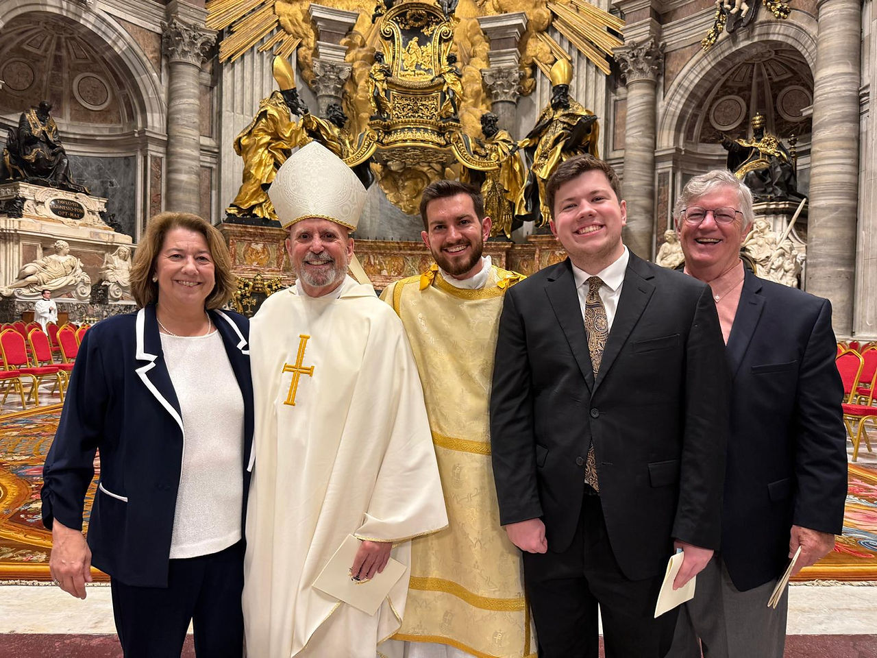 Smiling group of five posing in a grand ornate church, including two clergy in white and gold vestments, with red chairs behind.