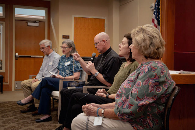 Five adults seated in a conference room, one man speaking into a microphone while others listen, with an American flag behind them.