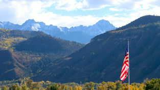 American flag on a pole in front of a mountain valley with autumn trees and snow-dusted peaks under a bright cloudy sky