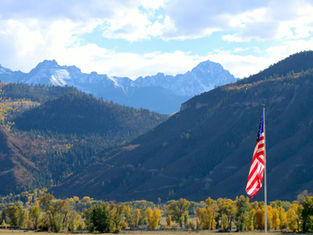American flag on a pole in front of a mountain valley with autumn trees and snow-dusted peaks under a bright cloudy sky