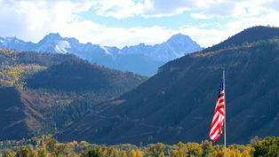 American flag on a pole in front of a mountain valley with autumn trees and snow-dusted peaks under a bright cloudy sky